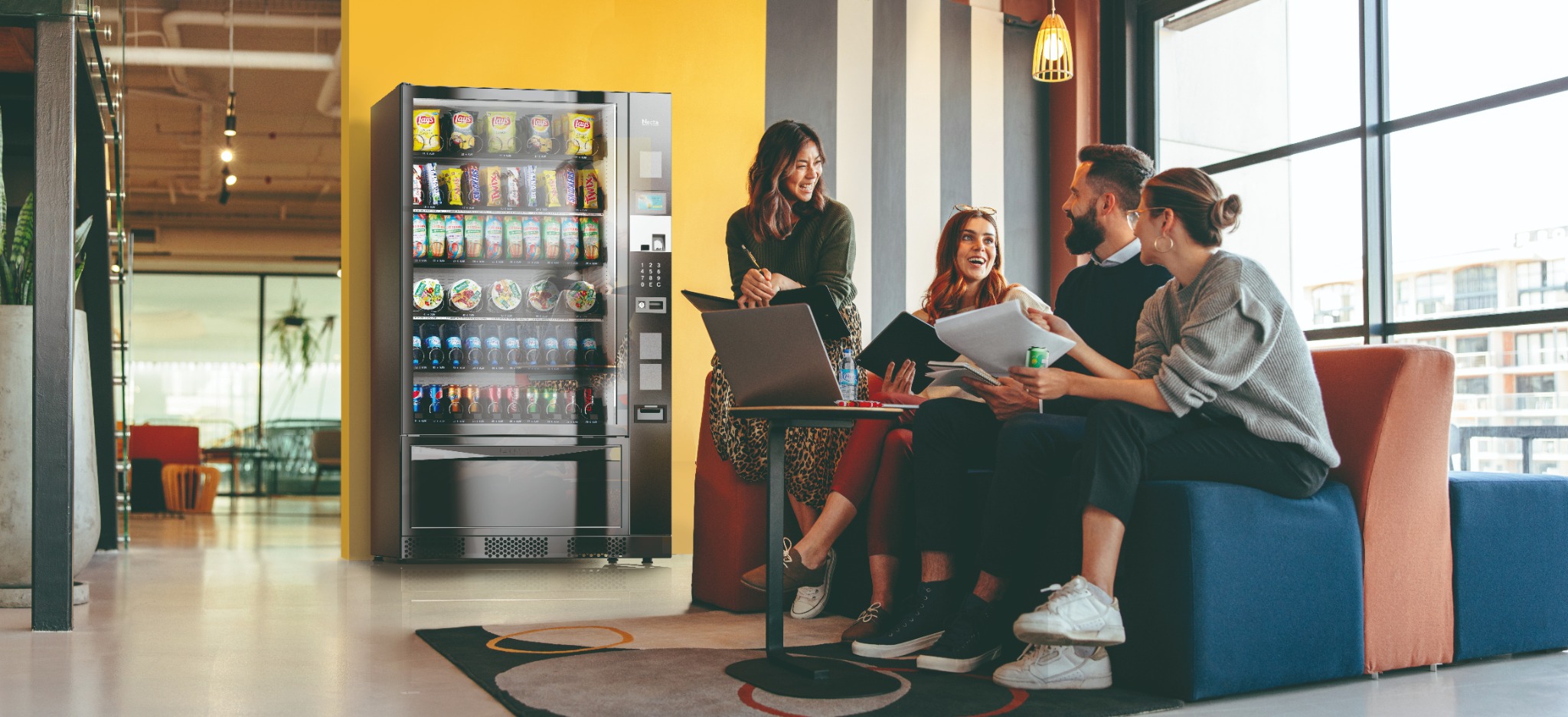 A snack and drink vending machine beside a team enjoying a break in a bright workplace
